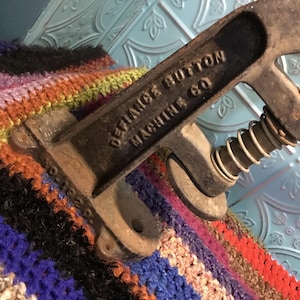 May include: Close-up of an antique, cast iron button machine with a spring mechanism. The machine is labeled "Defiance Button Machine Co."