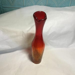 May include: A tall, slender, red glass vase with a flared top. The vase has a gradient color effect, transitioning from red at the top to orange and then to a brown base. The glass has a textured surface.