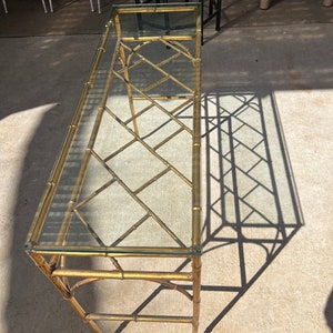 May include: A gold-toned bamboo-style console table with a glass top. The table has a diamond-shaped lattice design and is made of metal.
