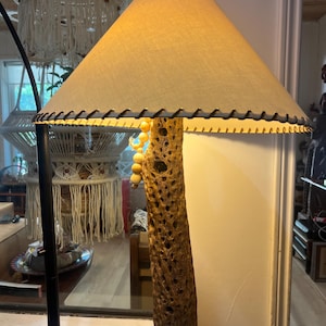 May include: A table lamp featuring a cylindrical base made from a textured, porous piece of cholla cactus wood. The lamp is topped with a conical, off-white fabric lampshade that has dark brown stitching along the bottom edge and light wooden beads.