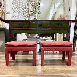 May include: Two red upholstered ottomans with a patterned design. The ottomans are rectangular with square legs. A dark green and brown wooden console table with three drawers is in the background.