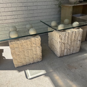 May include: A modern coffee table with a clear glass top supported by two textured, light-colored stone bases. The bases are rectangular and have a rough, carved appearance. The table is outdoors, with a brick wall in the background.
