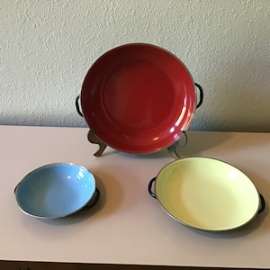 May include: Three enamelware bowls with black handles. The largest bowl is red, the medium bowl is yellow, and the smallest bowl is blue.