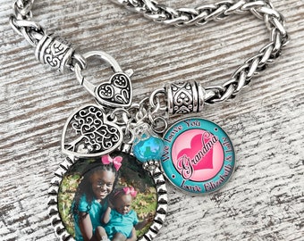 May include: Silver-toned charm bracelet with a braided chain. The bracelet features a heart-shaped clasp, a heart-shaped charm with a tree design, and a round charm with a photo of two people. A charm reads "We love you Grandma" with a pink heart.