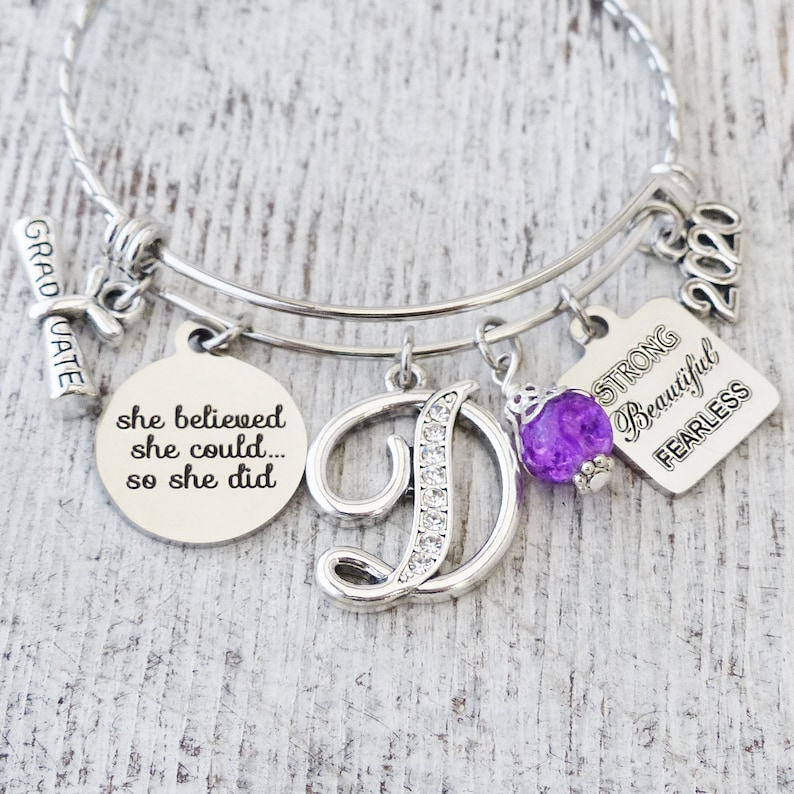May include: A silver charm bracelet with multiple charms, including a graduation cap, a letter D with rhinestones, a purple glass bead, and a square charm with the words "Strong, Beautiful, Fearless" and the year 2020. The charm with the letter D is the largest charm on the bracelet. The bracelet also has a round charm with the text "She believed she could... so she did."