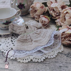 May include: A delicate, light brown doll's dress with a white lace trim. The dress is on a white crocheted doily.