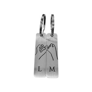 May include: Two silver keychains with a pinky promise design. Each keychain has a rectangular shape with a silver ring at the top. The keychains have the letters "L" and "M" printed on them.