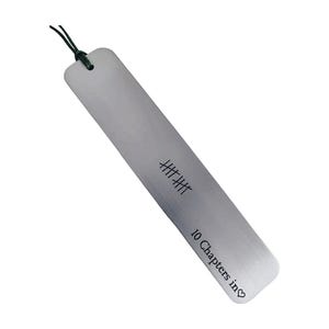 May include: A silver metal bookmark with a dark green string. The bookmark has etched markings and the text "10 Chapters in♡". The bookmark is rectangular with rounded edges.