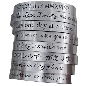 May include: A stack of silver bracelets with engraved inspirational messages, including "Love Fiercely", "Take one day at a time", "It begins with me", and "Forever in My Heart".