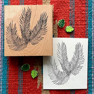 Puede incluir: Un sello de madera con un dibujo de línea negra de una hoja de palmera. El sello está sobre una tela de rayas rojas, naranjas y azules. Hay una imagen impresa de la hoja de palmera en una tarjeta blanca.