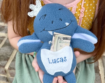 Könnte beinhalten: Ein blaues Plüschmonster mit weißem Bauch und einem grinsenden Gebiss. Das Monster hat eine Tasche auf dem Bauch, aus der ein Dollar-Schein herausragt. Die Tasche ist mit dem Namen "Lucas" bestickt.