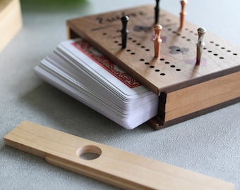 May include: Wooden cribbage board with a deck of cards inside. The board has holes for pegs and a sliding lid. The pegs are silver, copper, and black. The game is on a light gray surface.