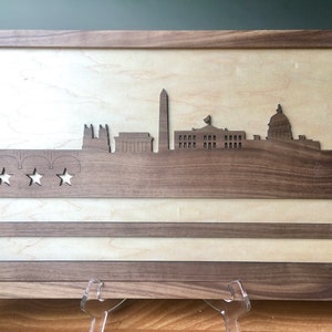 May include: A wooden framed artwork featuring a silhouette of the Washington D.C. skyline with three stars and stripes below. The artwork is made of walnut wood and has a light wood background.