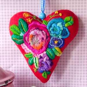 Op de afbeelding: Een rood vilten hartvormig ornament met geborduurde bloemen in tinten roze, blauw, groen en oranje. De bloemen zijn in een cluster gerangschikt en hebben een 3D-effect.