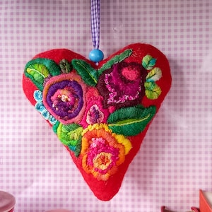 Op de afbeelding: Een rood vilten hartvormig ornament met geborduurde bloemen in tinten van roze, paars, groen en oranje. Het hart hangt aan een blauw-wit lint.
