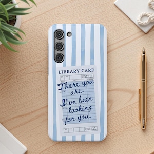 May include: A white phone case with blue vertical stripes and a library card design. The library card reads "There you are. I've been looking for you." A gold pen and a small plant are also visible.