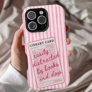 May include: Pink and white striped phone case with a library card design. The library card reads "easily distracted by books and dogs" in pink script. The case has a black camera module.
