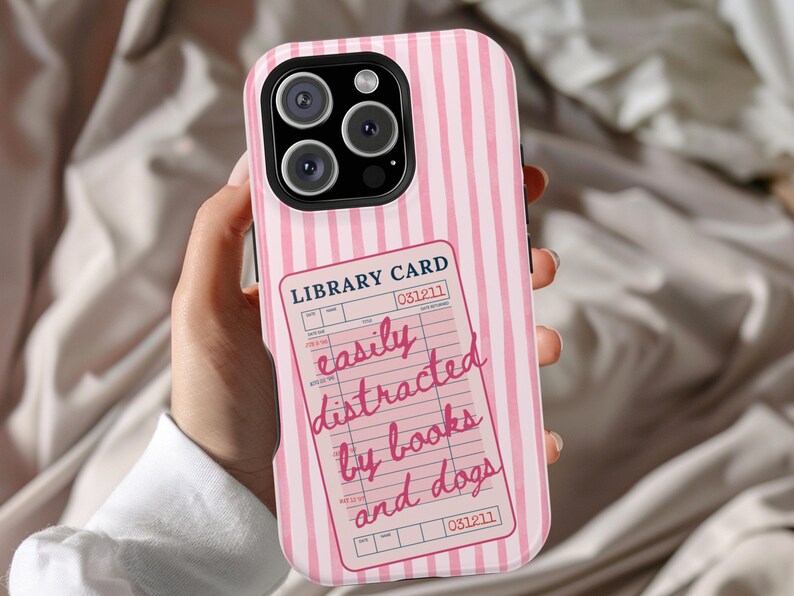 May include: Pink and white striped phone case with a library card design. The library card reads "easily distracted by books and dogs" in pink script. The case has a black camera module.