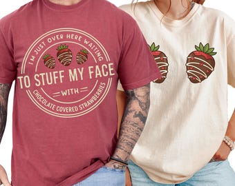 May include: Two t-shirts, one maroon and one cream-colored, with strawberry graphics. The maroon shirt has a circular design with text that reads "I'M JUST OVER HERE WAITING TO STUFF MY FACE WITH CHOCOLATE COVERED STRAWBERRIES." The cream shirt has two strawberry graphics.