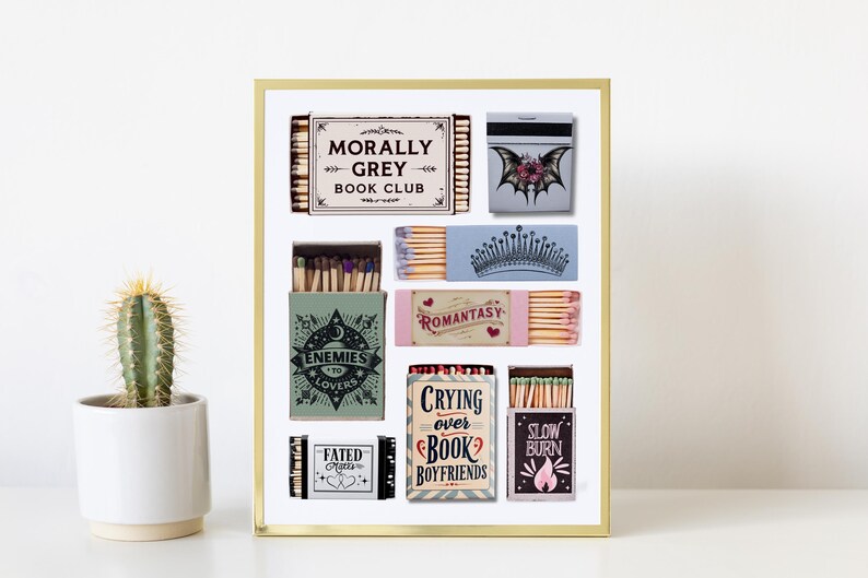 May include: A framed print featuring various matchbox designs with text like "Morally Grey Book Club" and "Enemies to Lovers." The matchboxes have different colors and graphics, including a crown and a bat. A cactus in a white pot is in the foreground.