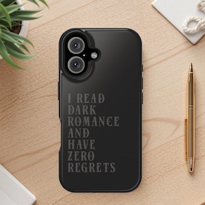 May include: Black phone case with the text "I READ DARK ROMANCE AND HAVE ZERO REGRETS". The case has a dual-lens camera cutout. A gold pen and a small potted plant are on a wooden surface.