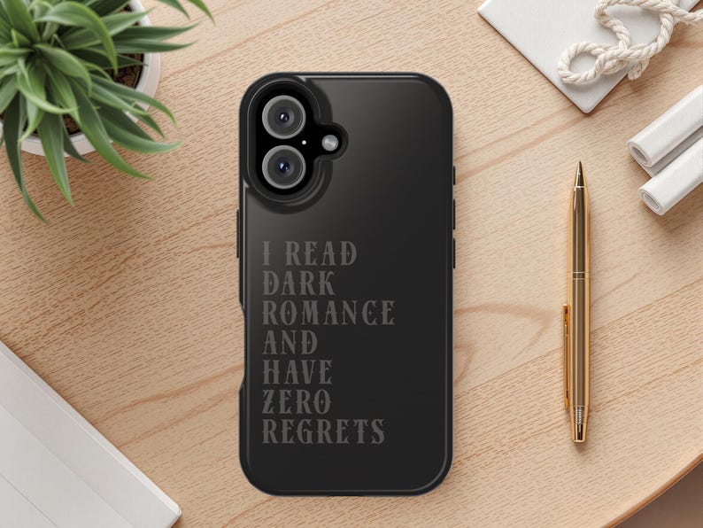 May include: Black phone case with the text "I READ DARK ROMANCE AND HAVE ZERO REGRETS". The case has a dual-lens camera cutout. A gold pen and a small potted plant are on a wooden surface.