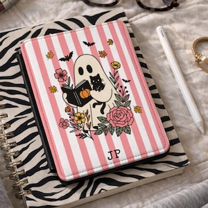 May include: A rectangular e-reader cover featuring a white and pink vertical striped background. The center graphic shows a ghost holding a book with a pumpkin illustration and a black cat, surrounded by colorful flowers, leaves, and bats. The initials JP are printed in black at the bottom.