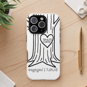 Op de afbeelding: Witte telefoonhoes met een zwart boomontwerp, een hartvormige gravure met de initialen "K&P", het woord "verloofd" en de datum "7.14.25".