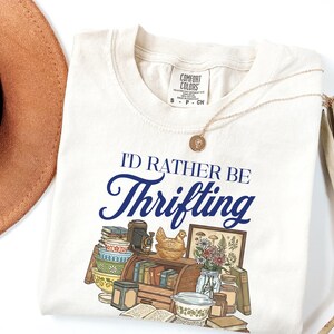 May include: Cream-colored t-shirt with the text "I'D RATHER BE Thrifting" in navy blue script. The shirt features a graphic of vintage items like books, a camera, and a chicken figurine. A gold necklace and a brown hat are also visible.