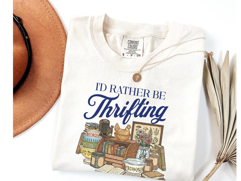 May include: Cream-colored t-shirt with the text "I'D RATHER BE Thrifting" in navy blue script. The shirt features a graphic of vintage items like books, a camera, and a chicken figurine. A gold necklace and a brown hat are also visible.