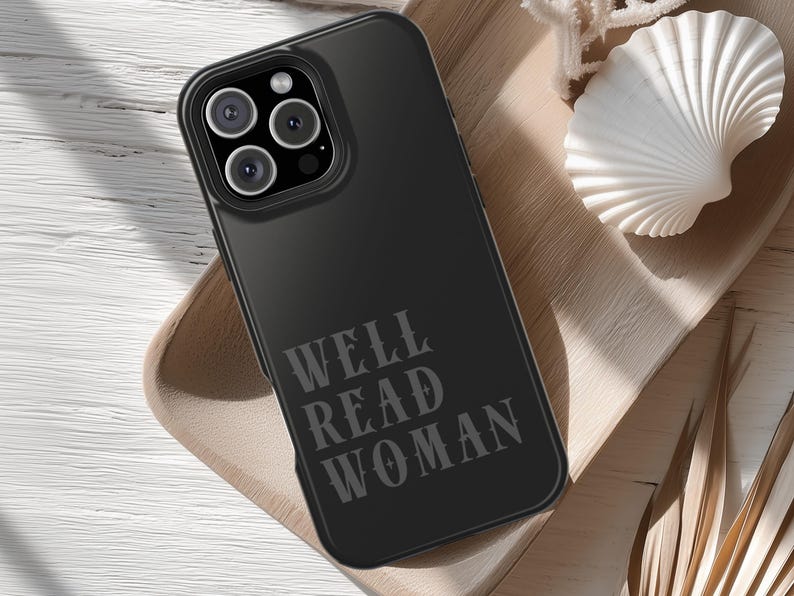 May include: Black phone case with the text "WELL READ WOMAN" in a serif font. The phone case has a black camera module with three lenses. The case is on a wooden tray with a white seashell.