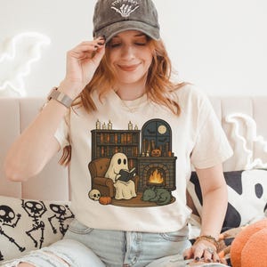 May include: Cream-colored t-shirt featuring a Halloween-themed illustration. The graphic depicts a ghost reading a book by a fireplace, with a bookshelf, pumpkin, and cat. The shirt is paired with light-wash jeans and a gray baseball cap.