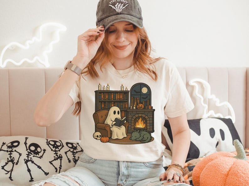 May include: Cream-colored t-shirt featuring a Halloween-themed illustration. The graphic depicts a ghost reading a book by a fireplace, with a bookshelf, pumpkin, and cat. The shirt is paired with light-wash jeans and a gray baseball cap.