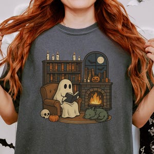 May include: Dark gray t-shirt featuring a cartoon ghost reading a book by a fireplace. The illustration includes a bookshelf, pumpkin, skull, sleeping dragon, and a window with a moon and bats. A cozy Halloween theme.