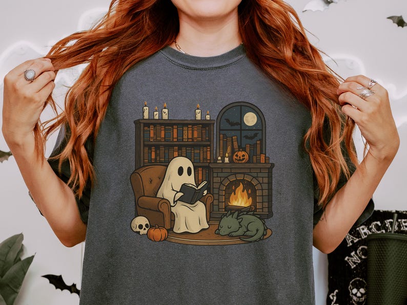 May include: Dark gray t-shirt featuring a cartoon ghost reading a book by a fireplace. The illustration includes a bookshelf, pumpkin, skull, sleeping dragon, and a window with a moon and bats. A cozy Halloween theme.