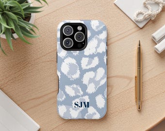 May include: A pale blue phone case featuring a white leopard print pattern. The case has a black camera module and navy blue initials "SJM". A gold pen and a small potted plant are also visible.