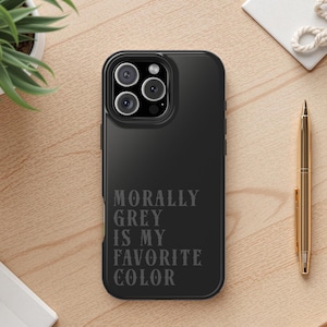 May include: A black phone case with the text "MORALLY GREY IS MY FAVORITE COLOR" in a stylized font. A gold pen and a small potted plant are on a wooden surface.