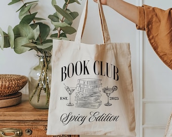 May include: Cream canvas tote bag featuring the text "BOOK CLUB Spicy Edition" in black, with illustrations of books and cocktails. The bag is being held up.