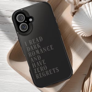 May include: Black phone case with the text "I READ DARK ROMANCE AND HAVE ZERO REGRETS" in a gray font. The phone case is on a wooden tray with a white seashell.