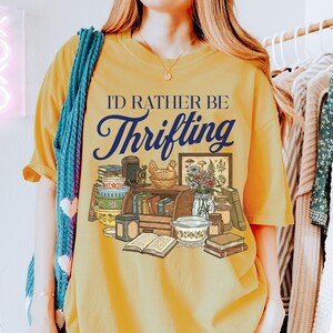 May include: Mustard yellow t-shirt with the text "I'd Rather Be Thrifting" in navy blue script. The graphic features a collection of vintage items, including books, bowls, and a framed picture. A blue scarf is draped over the shoulder.