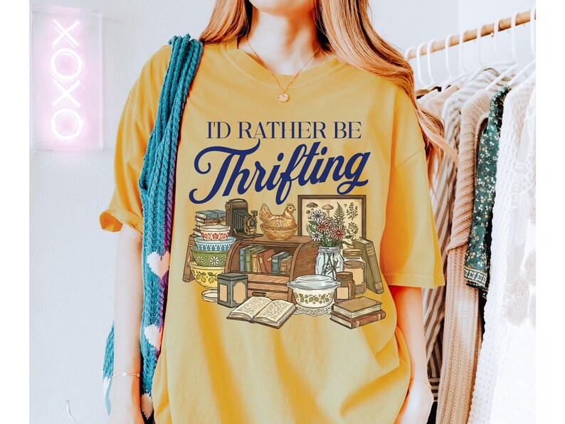 May include: Mustard yellow t-shirt with the text "I'd Rather Be Thrifting" in navy blue script. The graphic features a collection of vintage items, including books, bowls, and a framed picture. A blue scarf is draped over the shoulder.