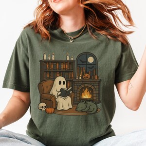 May include: Olive green t-shirt featuring a cartoon ghost reading a book in a cozy setting. The design includes a fireplace, bookshelf, pumpkin, skull, and a sleeping cat. A full moon and bats are visible through a window.