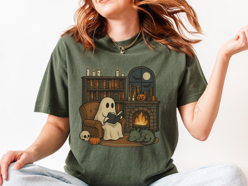May include: Olive green t-shirt featuring a cartoon ghost reading a book in a cozy setting. The design includes a fireplace, bookshelf, pumpkin, skull, and a sleeping cat. A full moon and bats are visible through a window.