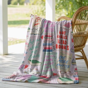 May include: A soft, patterned throw blanket draped over a wicker chair. The blanket features a repeating design of books, glasses, and hearts, with text that reads "Hot Girls Read Books" and "Ask me about my books."