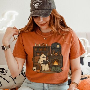 May include: Orange t-shirt featuring a Halloween-themed illustration. The design shows a ghost reading a book by a fireplace, with a cat, pumpkins, and a skull. The shirt also has a black cap with the text "Stay Spooky".