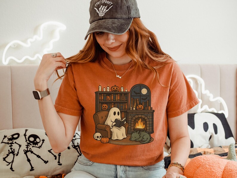 May include: Orange t-shirt featuring a Halloween-themed illustration. The design shows a ghost reading a book by a fireplace, with a cat, pumpkins, and a skull. The shirt also has a black cap with the text "Stay Spooky".