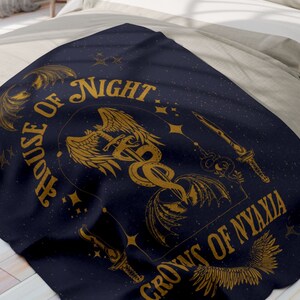 May include: A dark blue throw blanket with a gold design featuring a winged serpent, a sword, and the text "House of Night Crowns of Nyaxia".