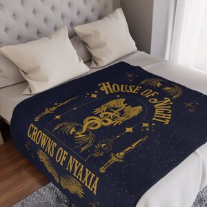 May include: A navy blue throw blanket with a gold design featuring a winged creature, a snake, and a sword. The text "House of Night" and "Crowns of Nyaxia" are written in gold lettering.