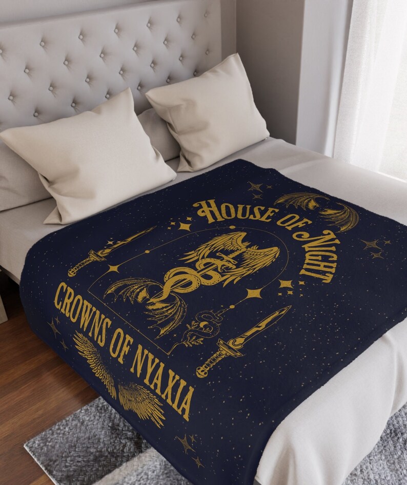May include: A navy blue throw blanket with a gold design featuring a winged creature, a snake, and a sword. The text "House of Night" and "Crowns of Nyaxia" are written in gold lettering.