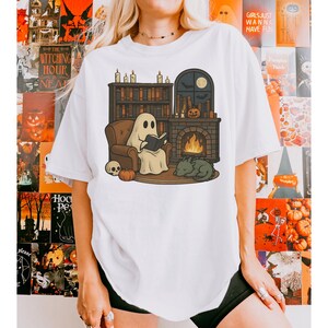 May include: White t-shirt featuring a cartoon ghost reading a book in front of a fireplace, with a cat, pumpkin, and skull. The scene is set in a cozy room with a bookshelf and a window with a moon. Halloween theme.